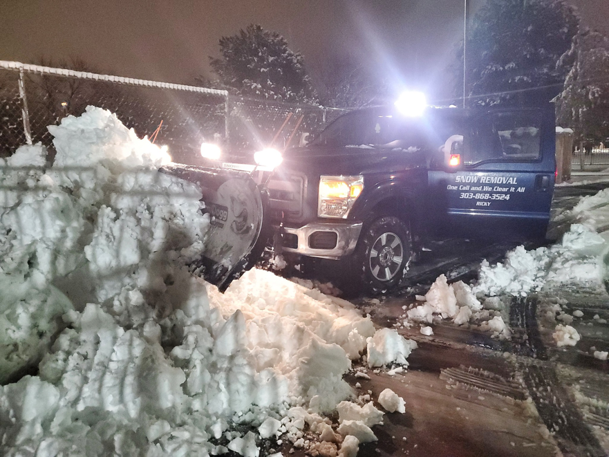A blue pickup truck with a snow plow actively clearing a large pile of snow at night for Ricky's Snow Removal in Springfield, VA.