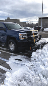 A blue pickup truck with a snow plow actively clearing snow from a parking lot by Denver Snow Removal in Denver, CO.