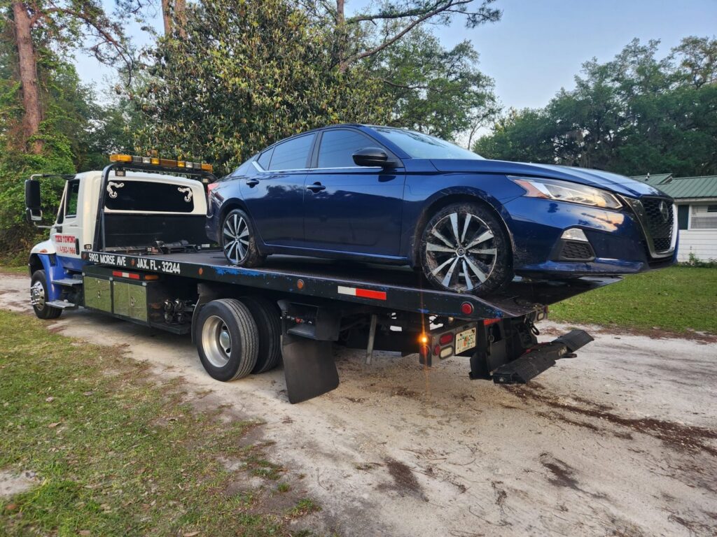 A blue sedan being unloaded from a flatbed tow truck by Tiki's Towing LLC in Jacksonville, FL.