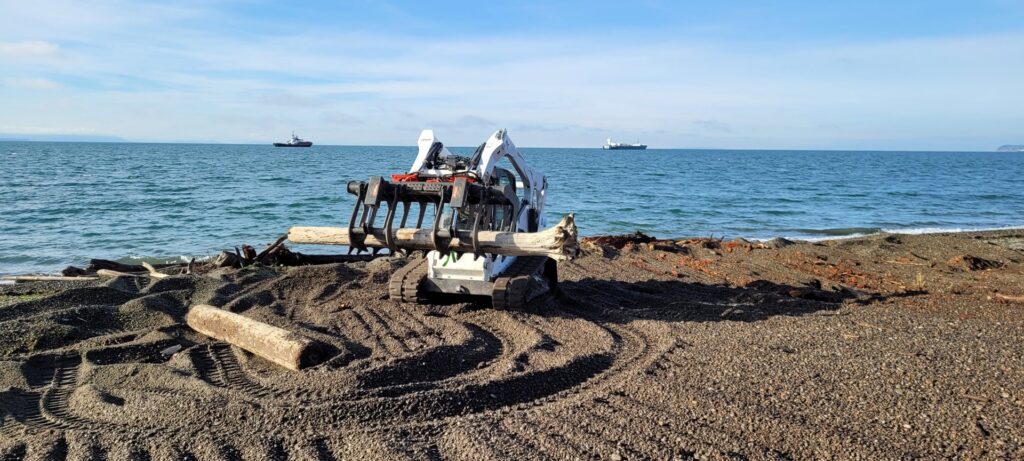 A Bobcat with a grapple attachment removing large logs from a beach, part of cleanup services by ROUGH 2 READY in Auburn, AL