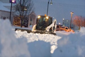 A Bobcat skid-steer loader with a plow attachment clearing snow from a commercial property at night for JC Snow Management in Mount Vernon, NY