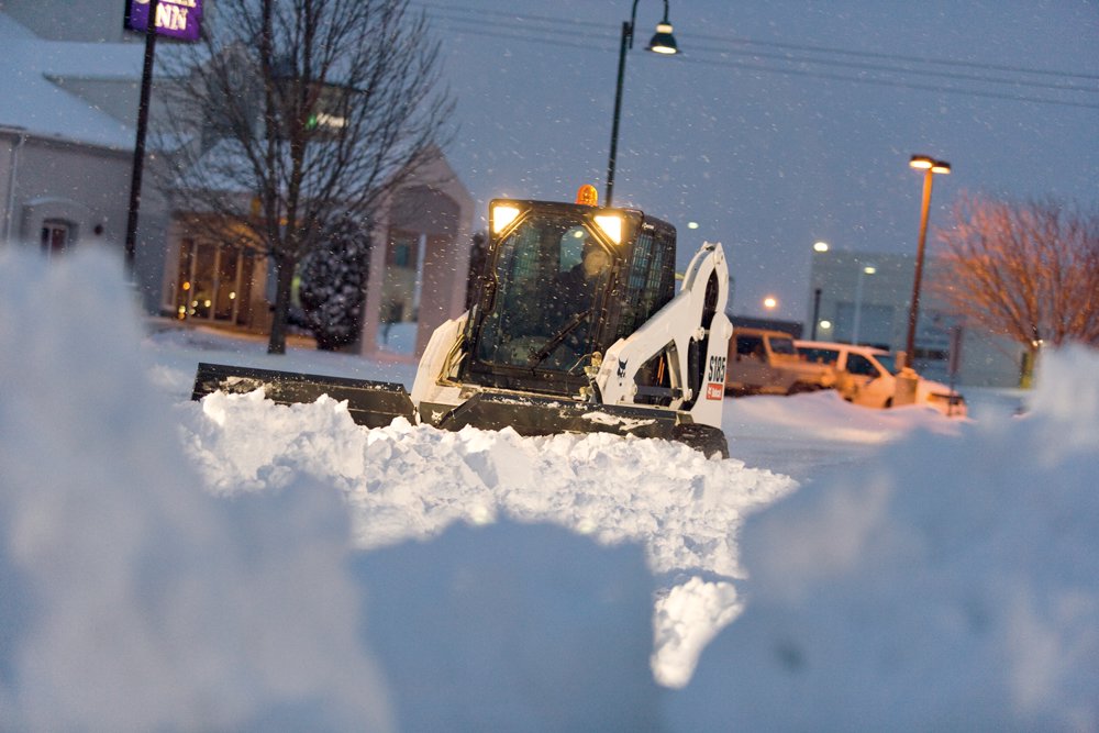 A Bobcat skid-steer loader with a plow attachment clearing snow from a commercial property at night for JC Snow Management in Mount Vernon, NY