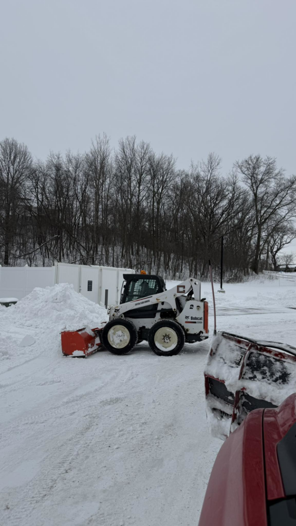 A Bobcat skid steer with a plow attachment actively clearing snow in a large area by Alex's lawn care in Rochester, MN.