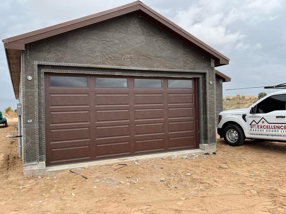 A brown panel garage door installed on a new construction home by Excellence Garage Doors LLC in Albuquerque, NM, with a service truck visible.