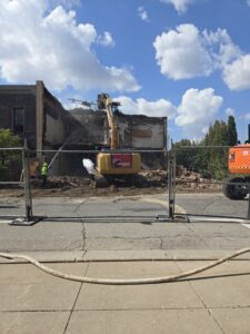 A building undergoing demolition by an excavator from DeCarlo Demolition Company, seen behind a fence in Des Moines, IA.