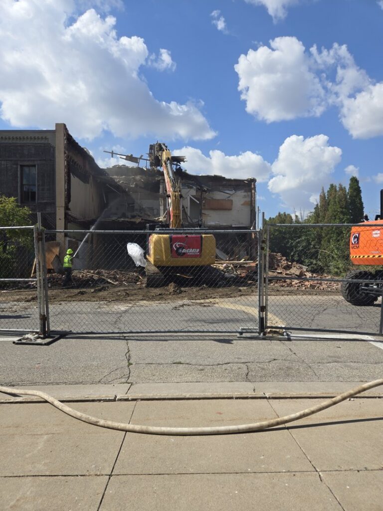 A building undergoing demolition by an excavator from DeCarlo Demolition Company, seen behind a fence in Des Moines, IA.