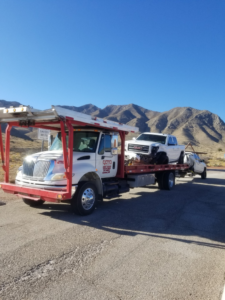 A car carrier tow truck from Gama Towing LLC transporting multiple vehicles on a road with mountains in Carlsbad, NM.