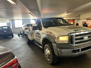 A white car being towed by a wheel lift tow truck from ABOD Towing LLC in New Orleans, LA, inside a parking garage.