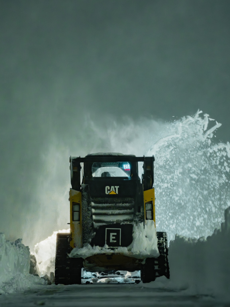 A yellow CAT skid steer with a snow blower attachment actively clearing snow for Elcor Construction in Rochester, MN.