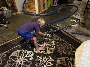 A child operating a carpet cleaning wand on a patterned rug, learning the trade at FM Cleaning in West Fargo, ND.
