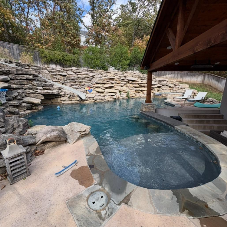 A sparkling clean backyard pool with rock features and a cleaning brush, maintained by AW Pool Service in Tulsa, OK.
