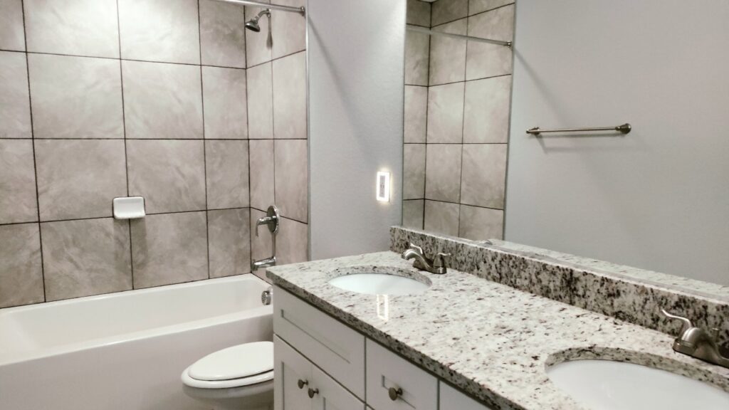 A clean bathroom with a double vanity, granite countertops, and tiled shower, expertly cleaned by Tessa's Cleaning Service in Chesapeake, VA.