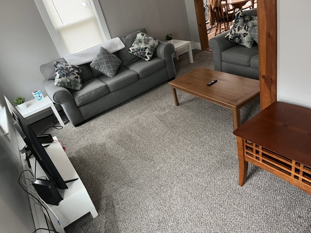 A clean living room featuring a grey sofa, coffee table, and vacuumed carpet, maintained by Make It Shine in Worcester, MA