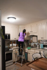A professional cleaner on a step stool wiping down kitchen cabinets for Sunshine Cleaning & Painting in Raleigh, NC.