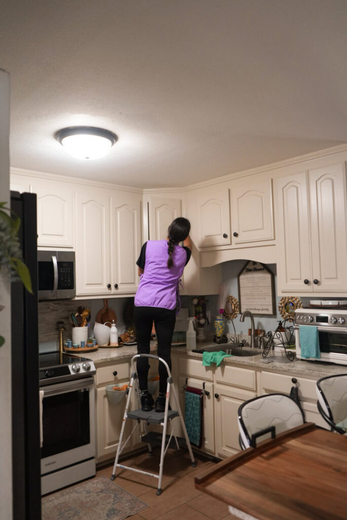 A professional cleaner on a step stool wiping down kitchen cabinets for Sunshine Cleaning & Painting in Raleigh, NC.