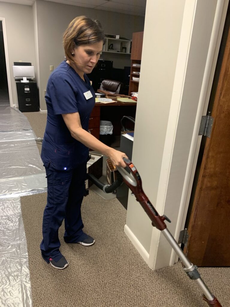 A professional cleaner in blue scrubs vacuuming a carpeted hallway, showcasing cleaning services by Precise Cleaning Solutions in Auburn, AL.