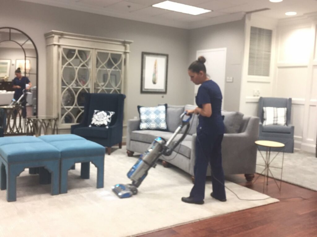 A professional cleaner in blue scrubs vacuuming a rug in a modern living room, demonstrating house cleaning by Precise Cleaning Solutions in Auburn, AL.