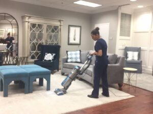 A professional cleaner in blue scrubs vacuuming a rug in a modern living room, demonstrating house cleaning by Precise Cleaning Solutions in Auburn, AL.