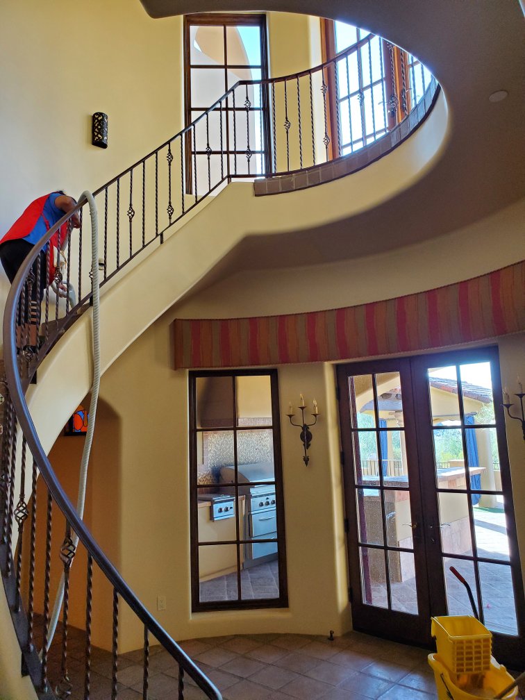 A house cleaner vacuuming a spiral staircase with a hose attachment for Arizona Housekeeping Service in Phoenix, AZ.