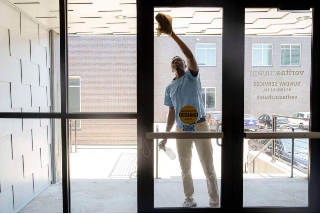 A cleaner from MJ Commercial & Residential Cleaning, LLC washing a glass door with spray and cloth in Cedar Rapids, IA.