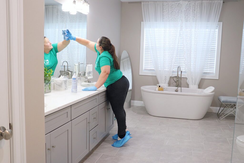 A house cleaner from Supreme Home Cleaning, Inc wiping a bathroom mirror, wearing protective shoe covers in Boston, MA.
