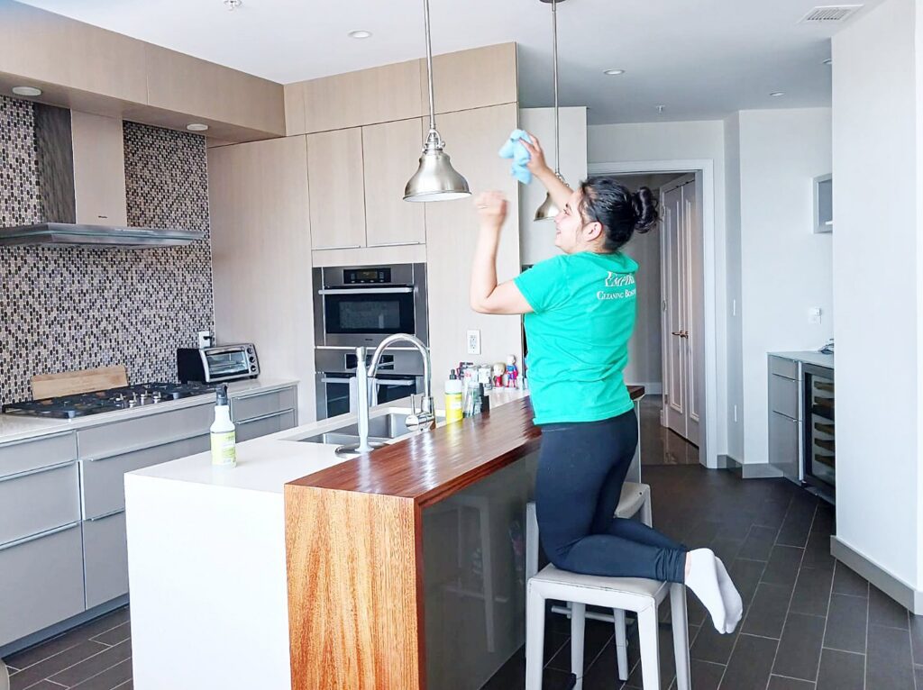 A house cleaner from Supreme Home Cleaning, Inc wiping kitchen light fixtures while standing on a stool in Boston, MA.