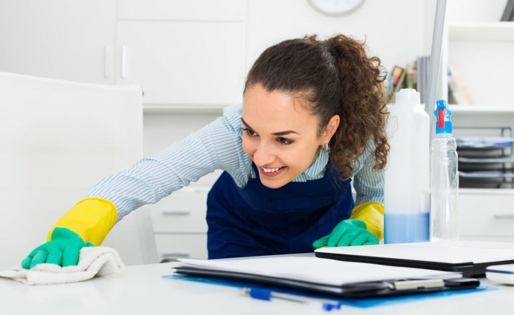 A smiling house cleaner in an apron and gloves wiping down an office desk for Mariahs House cleaning in Renton, WA.