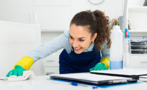 A smiling house cleaner in an apron and gloves wiping down an office desk for Mariahs House cleaning in Renton, WA.