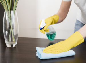 A cleaner wearing yellow gloves spraying and wiping a wooden table surface for PAP-Cleaning Services in Fort Lauderdale, FL.