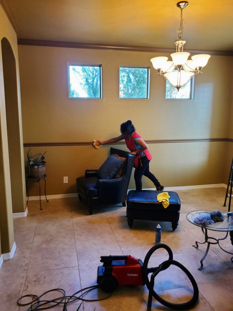 A house cleaner wiping a wall or furniture in a living room with a vacuum nearby for Arizona Housekeeping Service in Phoenix, AZ.