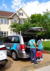 Two professional cleaners from Cozy Cleaning VA unloading cleaning supplies from their branded van in front of a client's home in Richmond, VA.