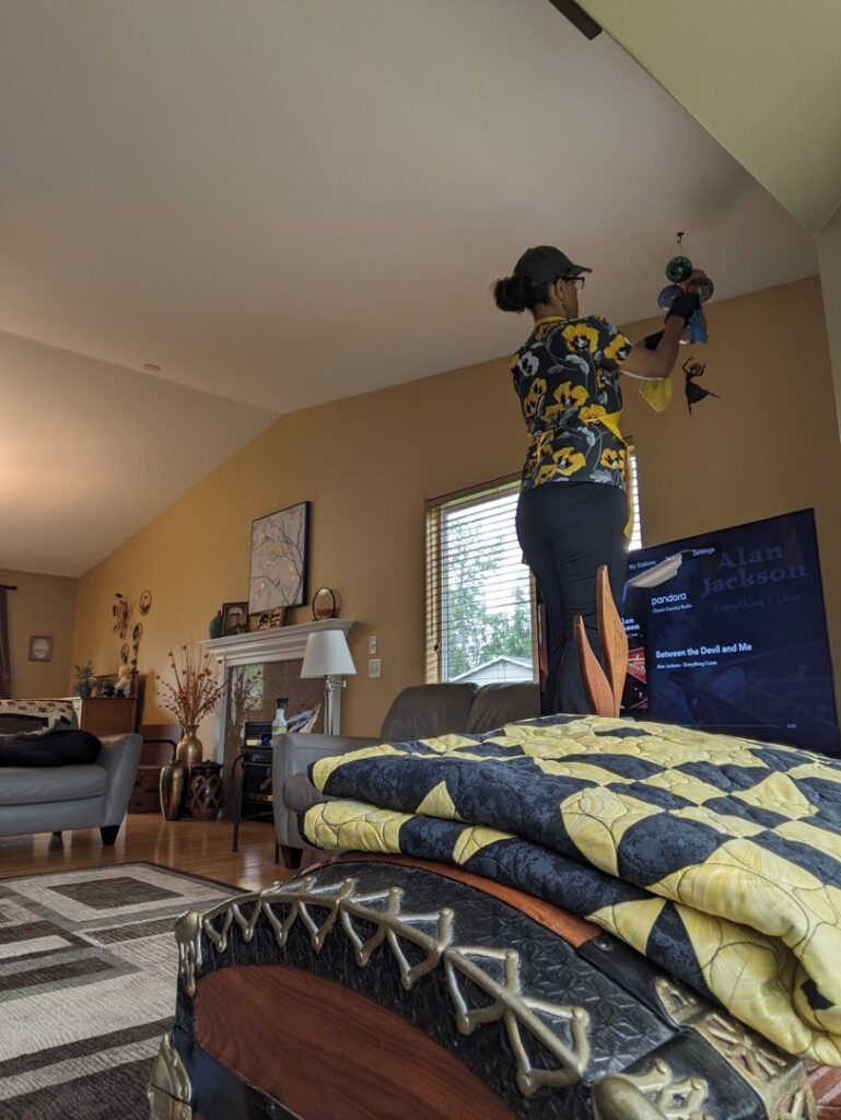 A house cleaner from Leave The Dust To Me Cleaning Services LLC in Anchorage, AK, cleaning a high ceiling fan in a living room.
