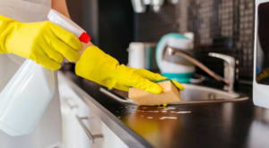 A person wearing yellow gloves sprays and scrubs a kitchen countertop, demonstrating house cleaning services by The Cathartic Couple in Cheyenne, WY.