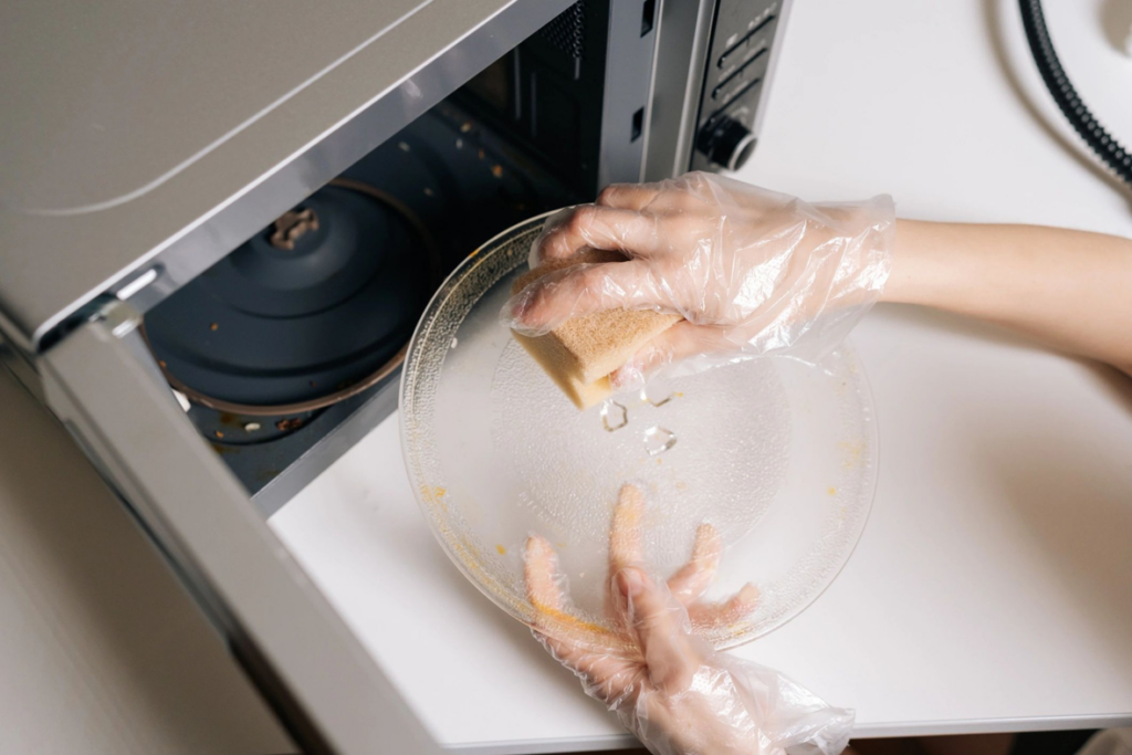 A person wearing gloves cleaning a microwave turntable with a sponge, demonstrating a house cleaning service by Two Gals & A Mop, LLC in Mobile, AL.