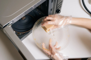A person wearing gloves cleaning a microwave turntable with a sponge, demonstrating a house cleaning service by Two Gals & A Mop, LLC in Mobile, AL.