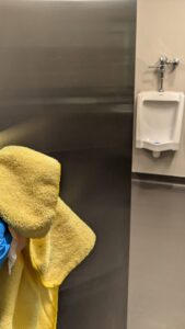 A professional cleaner from Leave The Dust To Me Cleaning Services LLC in Anchorage, AK, cleaning a stainless steel surface next to a urinal.