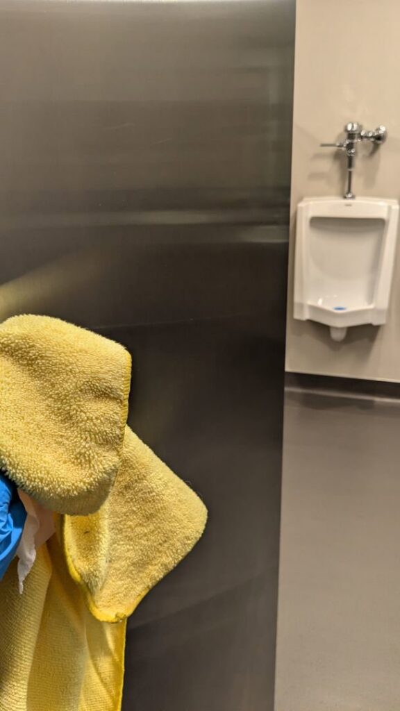 A professional cleaner from Leave The Dust To Me Cleaning Services LLC in Anchorage, AK, cleaning a stainless steel surface next to a urinal.