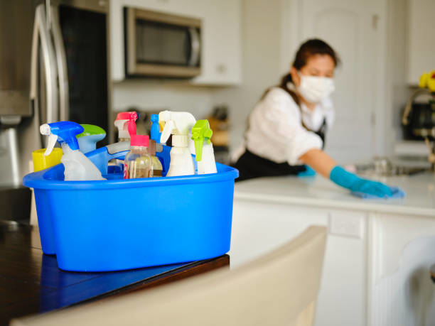 Cleaning supplies in a caddy, with a cleaner wiping a kitchen counter in the background for Clara's Cleaning LLC in Oklahoma City, OK.
