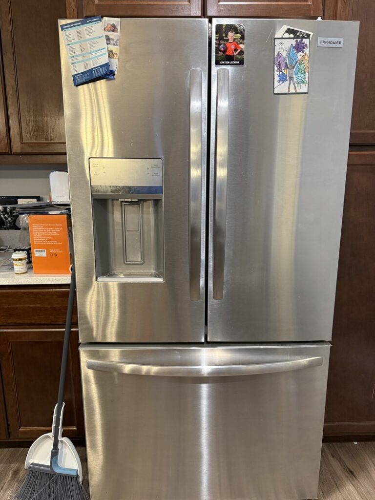 A broom and dustpan placed next to a stainless steel refrigerator, indicating cleaning work by Messy To Clean in Aurora, IL.