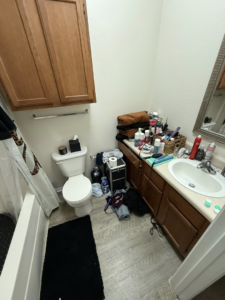 A cluttered bathroom with many items on the counter and floor, before house cleaning by WWJM Cleaning Service in Oklahoma City, OK.