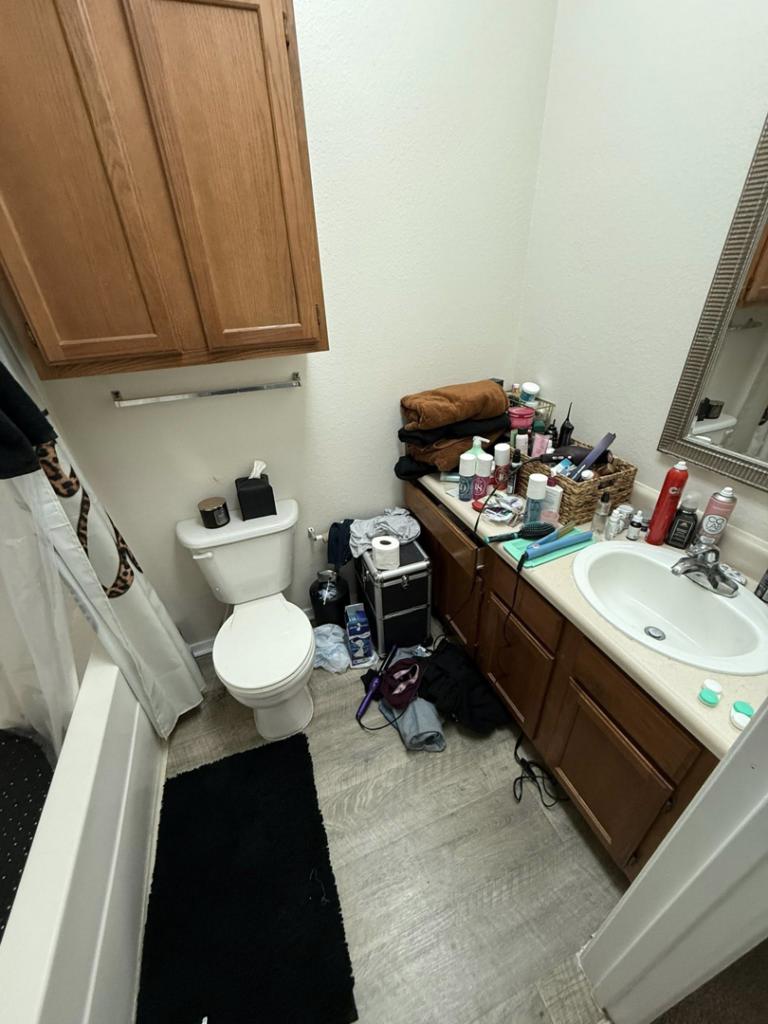 A cluttered bathroom with many items on the counter and floor, before house cleaning by WWJM Cleaning Service in Oklahoma City, OK.