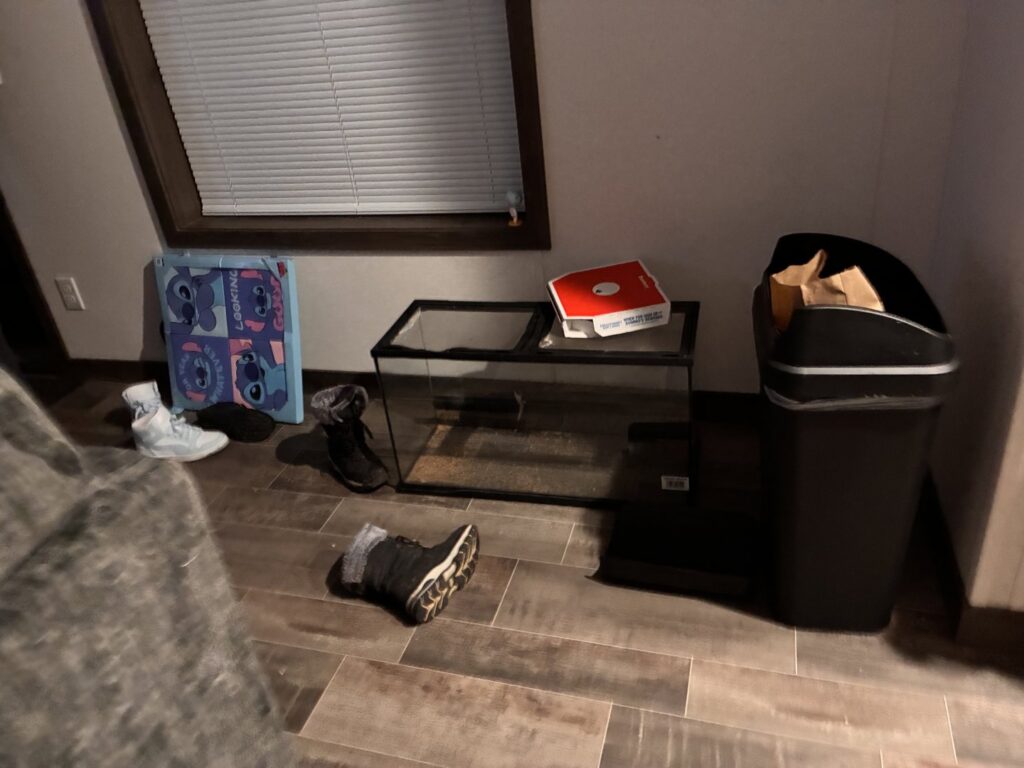 A cluttered floor with shoes, an empty fish tank, and a trash can, awaiting cleaning by Heavenly Harlots Maid Cleaning Services in Grand Forks, ND.