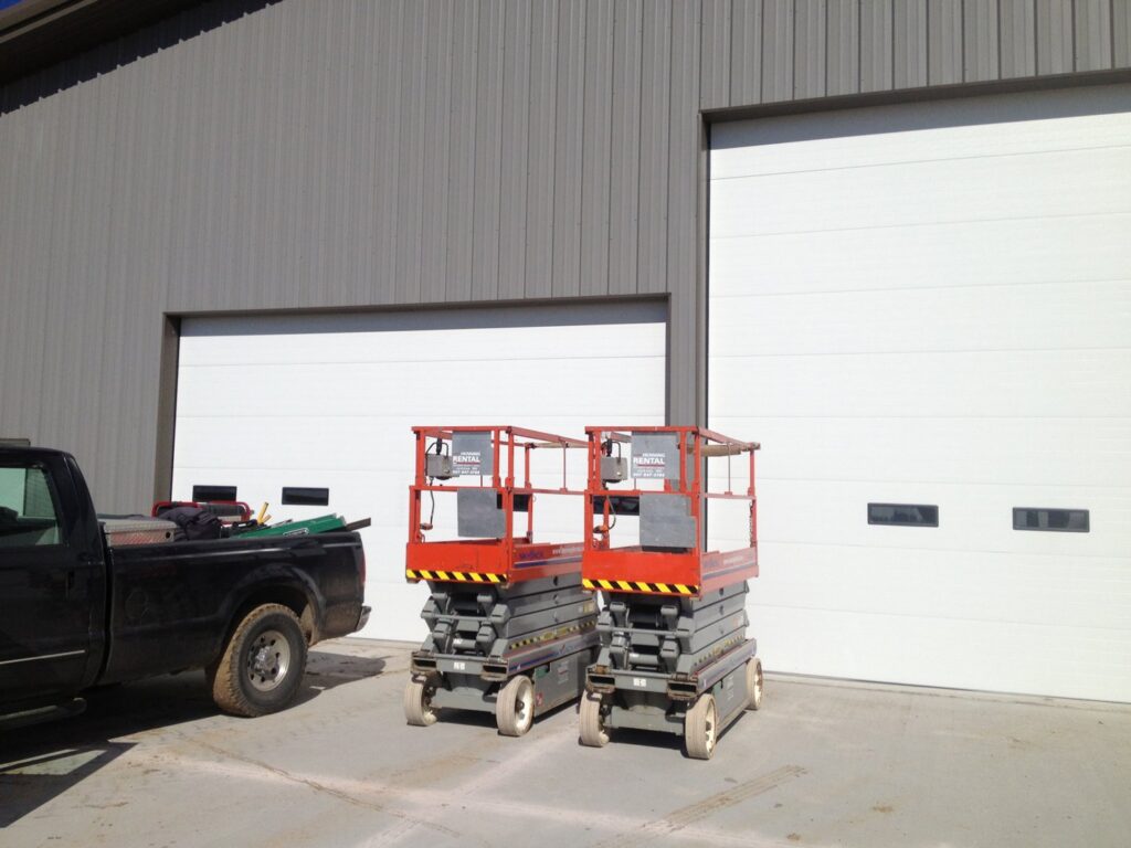 Two large white commercial garage doors installed on a grey metal building with scissor lifts nearby by Alberts Custom Door Company in Wichita, KS.