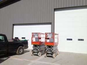 Two large white commercial garage doors installed on a grey metal building with scissor lifts nearby by Alberts Custom Door Company in Wichita, KS.