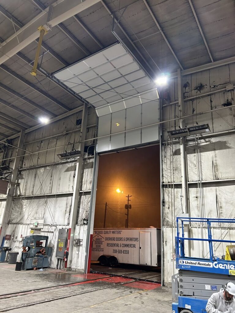 A commercial garage door partially open with a Barrett Overhead Doors branded trailer on site in Idaho Falls, ID.