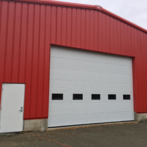 A large white commercial overhead garage door installed on a red corrugated building by American Dock & Door Co. in Warwick, RI.