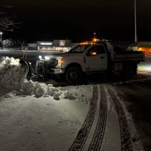 A CJS Landscape truck with a snow plow clearing a commercial parking lot at night in Green Bay, WI.