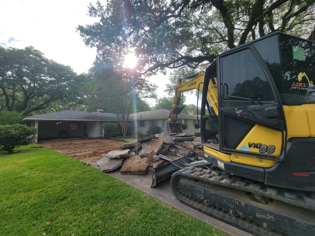 An excavator from Judice Dirt & Demolition breaking up a concrete driveway in front of a house in Lafayette, LA.