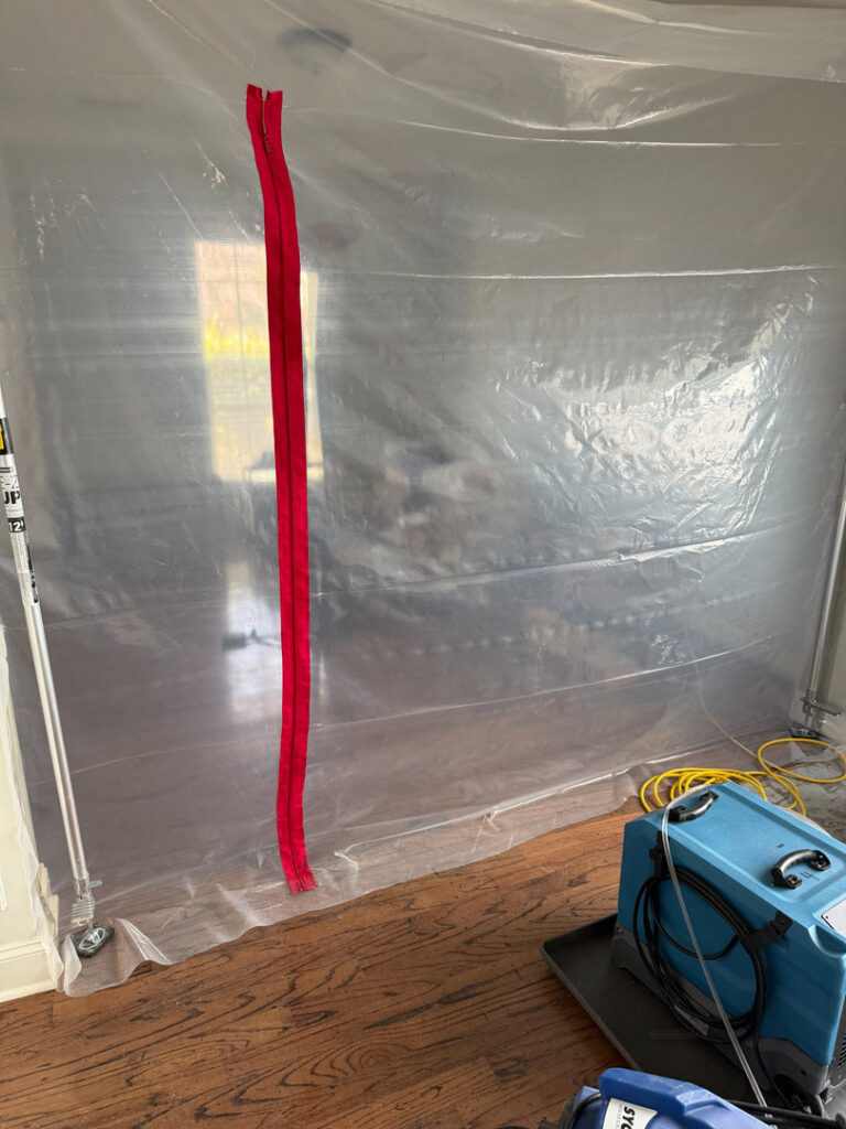 A room sealed with plastic containment and a red zipper, with an air mover for water damage drying by Triangle Restoration in Raleigh, NC.