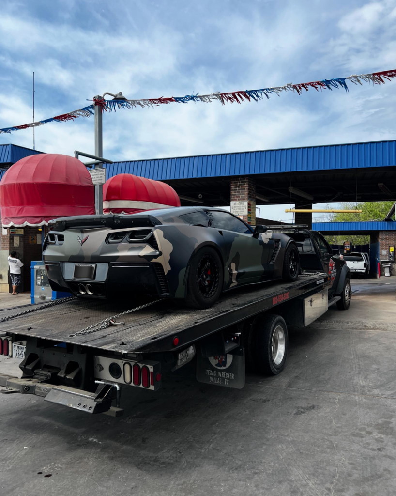 A camouflage Corvette being transported on a flatbed tow truck by Heavy Hookz Towing in Fort Worth, TX.